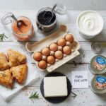 An overhead flat lay showcasing various budget-friendly protein options including cooked chicken, a carton of eggs, jars of lentils and black beans, plain yogurt, tofu, and canned tuna, with a note "FUEL FOR LESS" on a rustic white wooden table.