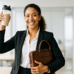Smiling professional woman in a black suit holding a protein shake bottle and a brown leather briefcase in a modern office setting.