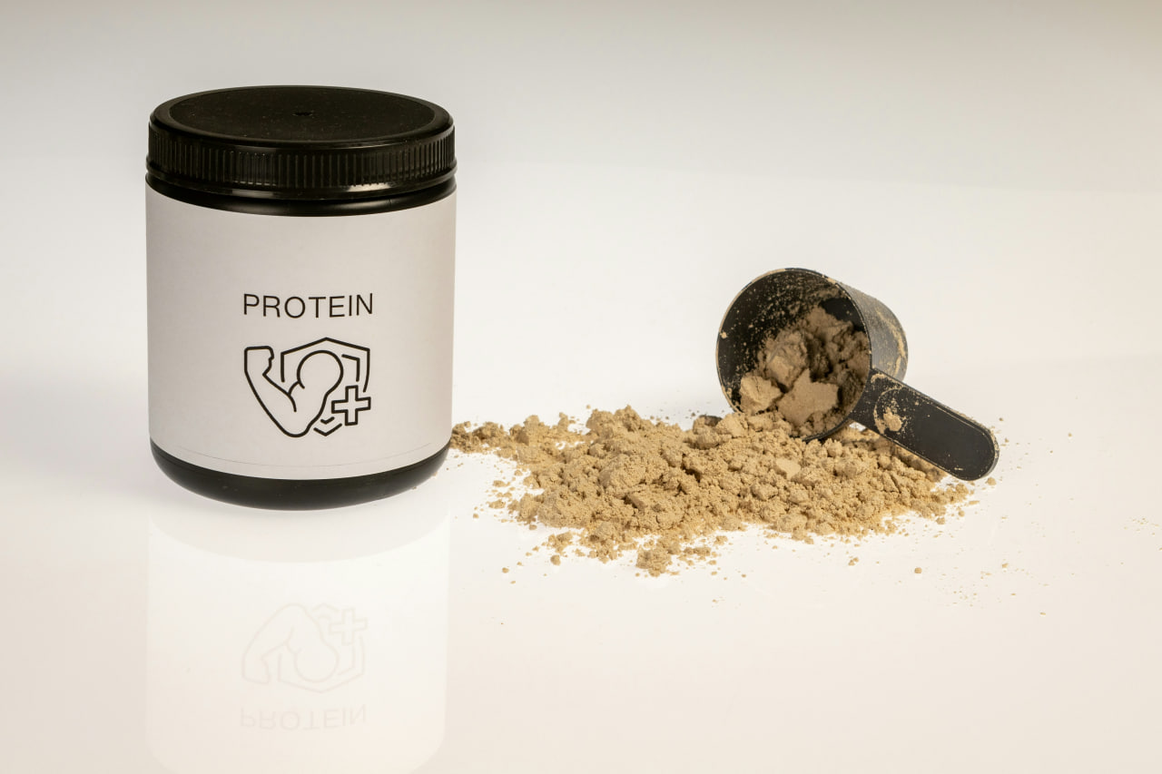 Protein powder for fat loss: A tub of protein powder with a flexing arm icon, alongside a scoop filled with brown powder on a white surface, representing a key supplement for weight management.